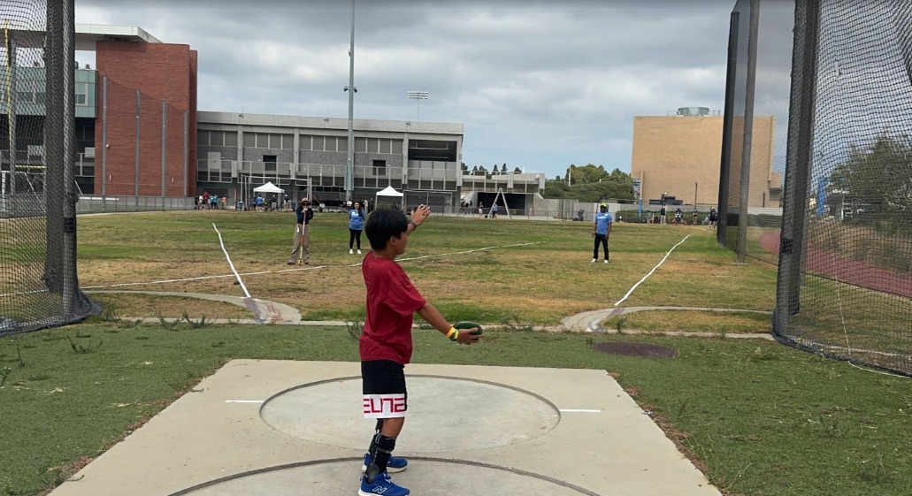 Ryle standing in the discus circle inside the cage, arm back, mid-throw at a meet