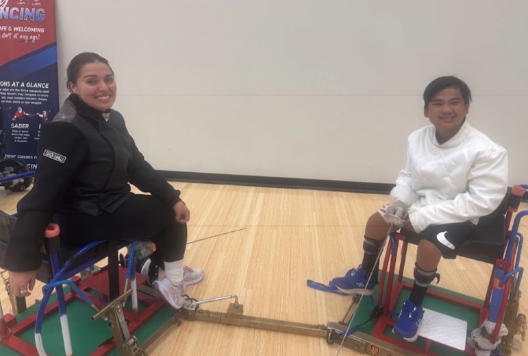 Ryle in fencing whites seated in his fencing frame, smiling at his opponent across the strip after their bout