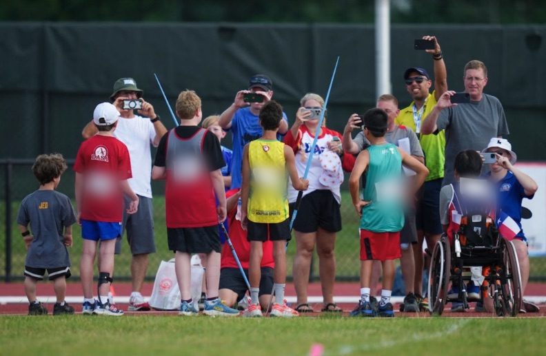 A crowd of young athletes, parents and coaches packed around the javelin runway, every one of them filming