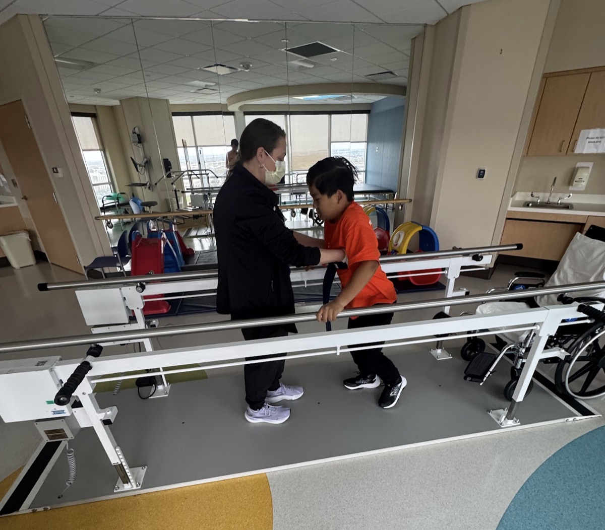 Ryle walking between parallel bars with mom's help during physical therapy